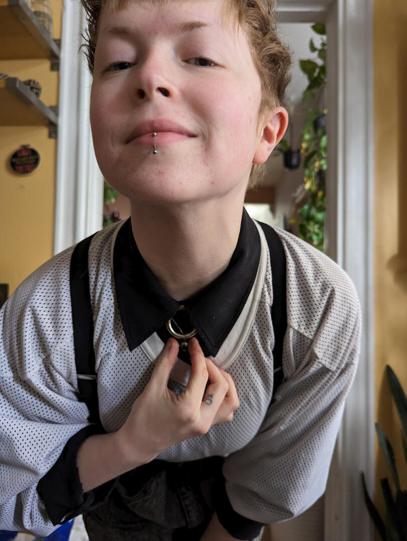 Atlas Katari, a blonde androgynous person, looks up and to the left of the camera with their head on their fist.